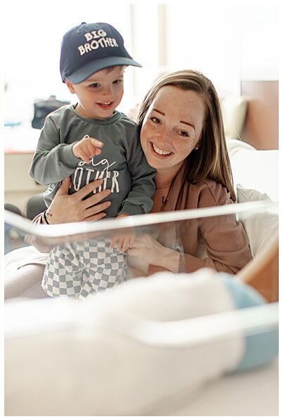 Mother holding toddler son, both are smiling looking at the newborn brother. Toddler is pointing happily at newborn