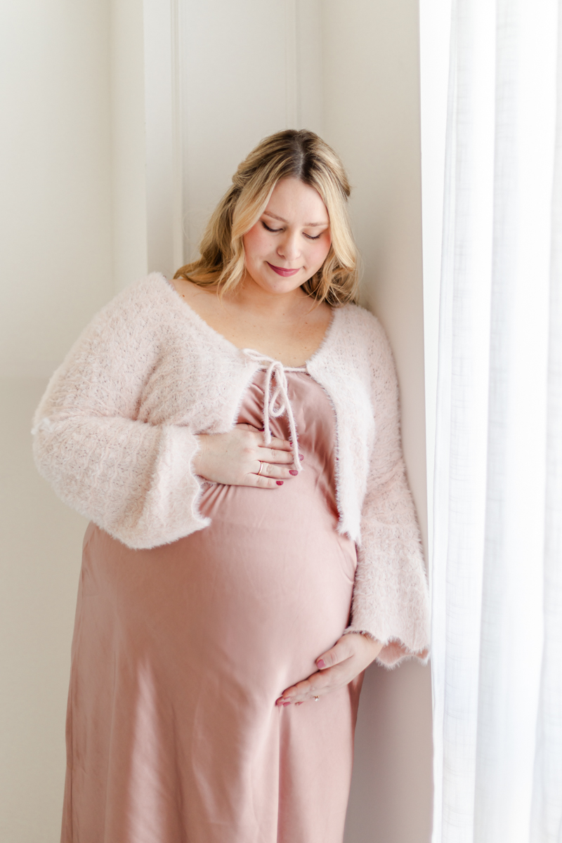 Mother in light pink silk dress and pink knit cardigan, cupping her baby bump, leaning against a window, gazing at baby bump.