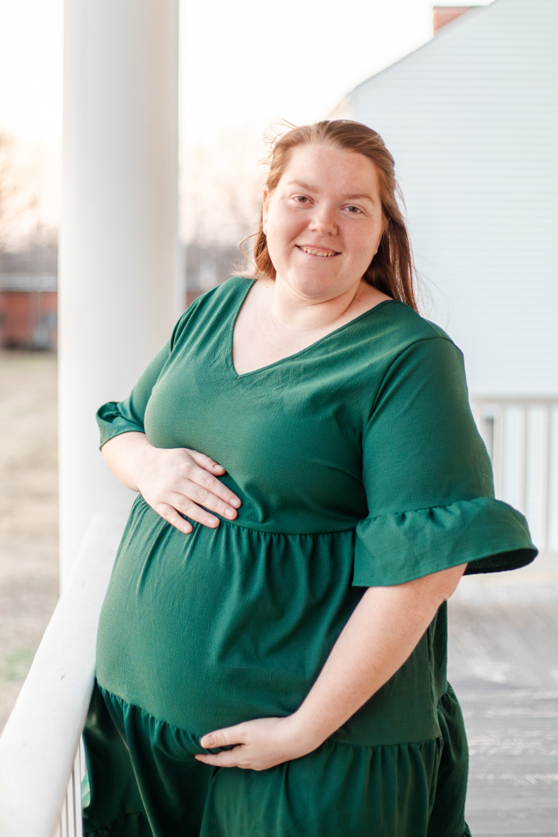 mother holding maternity belly, one hand on top and one on bottom, she is smiling into the camera on a white columned porch.