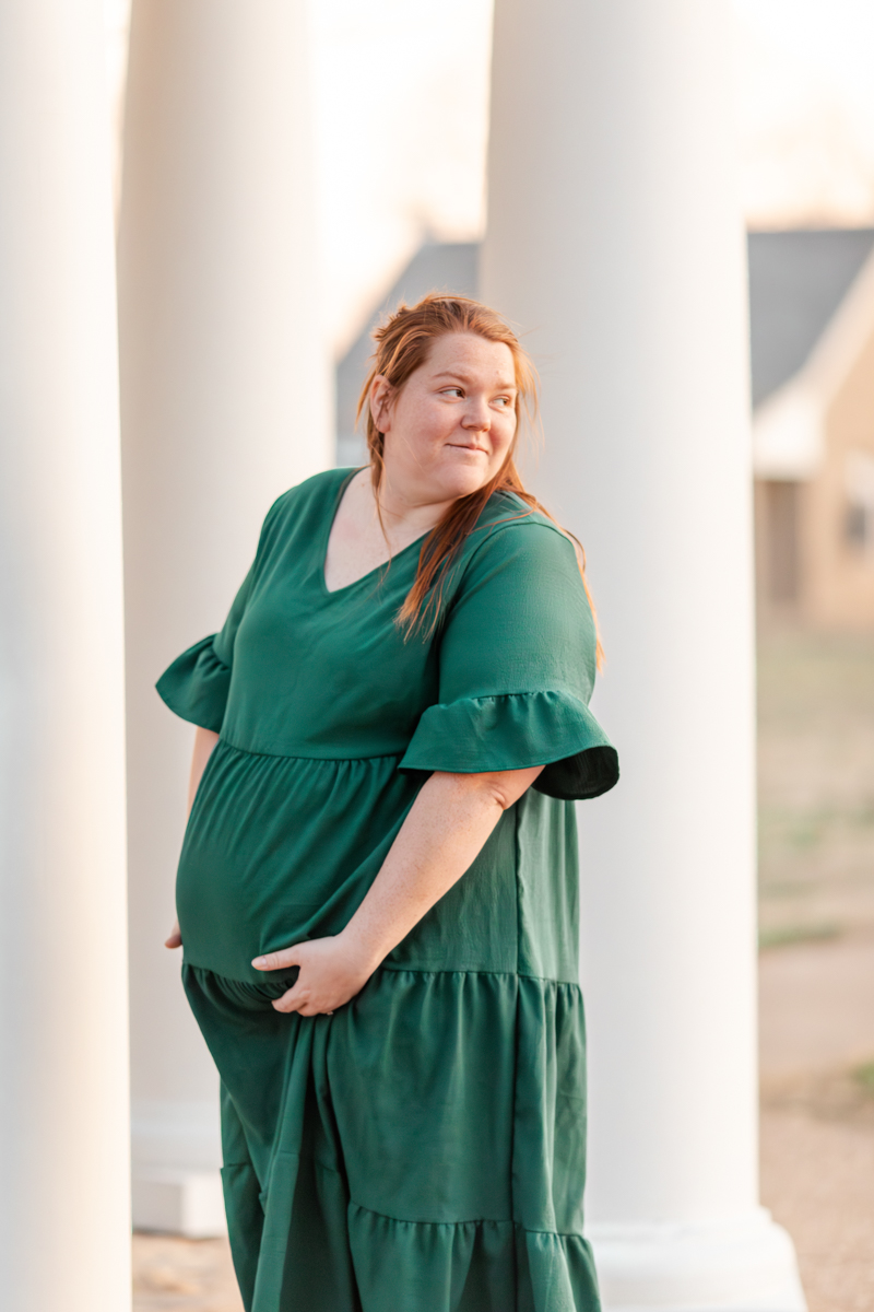 Pregnant mother in a long green dress, tenderly holding baby bump and looking over her shoulder off camera, standing in front of tall white pillars