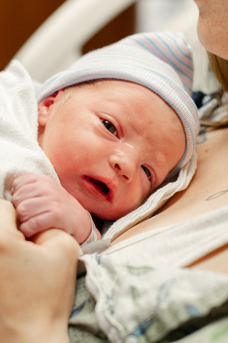 Newborn baby resting on mother's chest, holding her thumb, with a peaceful expression.
