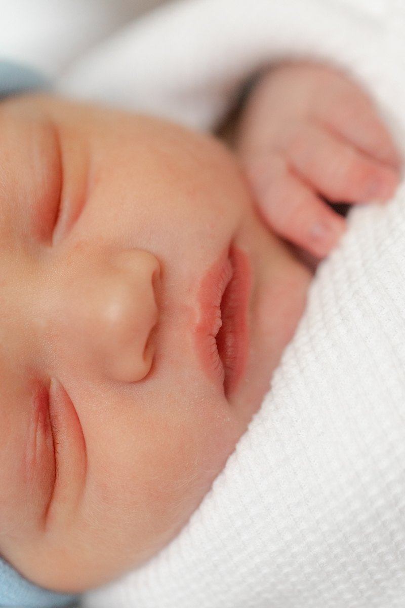 First 48 image of newborn baby boy, swaddled in hospital white blanket and blue cap. Close up of his facial features