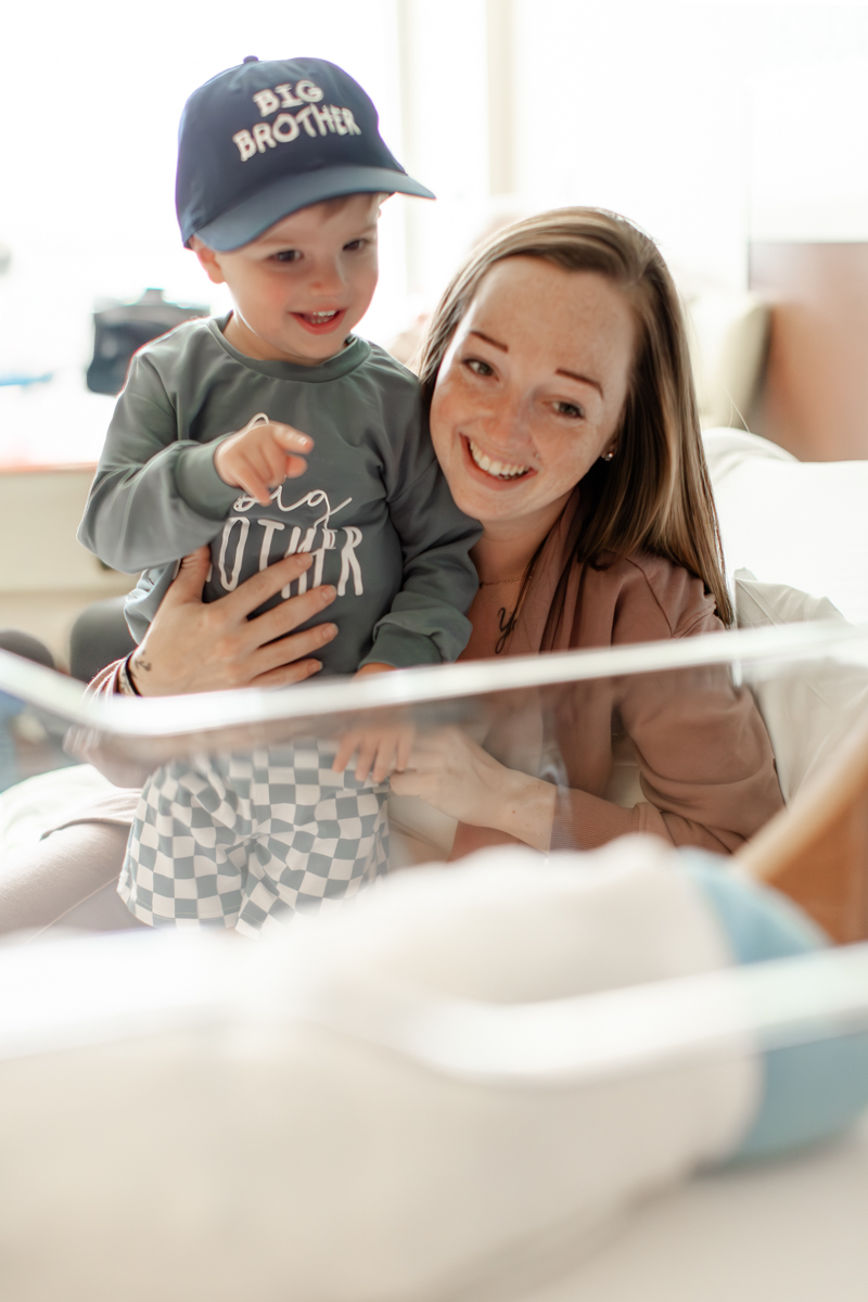 Mother holding toddler son, both are smiling looking at the newborn brother. Toddler is pointing happily at newborn