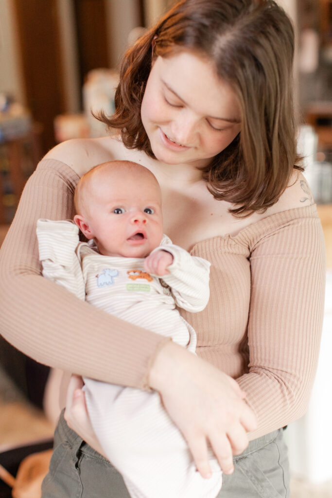 Mother holding and looking down at newborn while holding him to her chest. Baby boy is looking at the camera with wide eyes and a baggy sleeper.