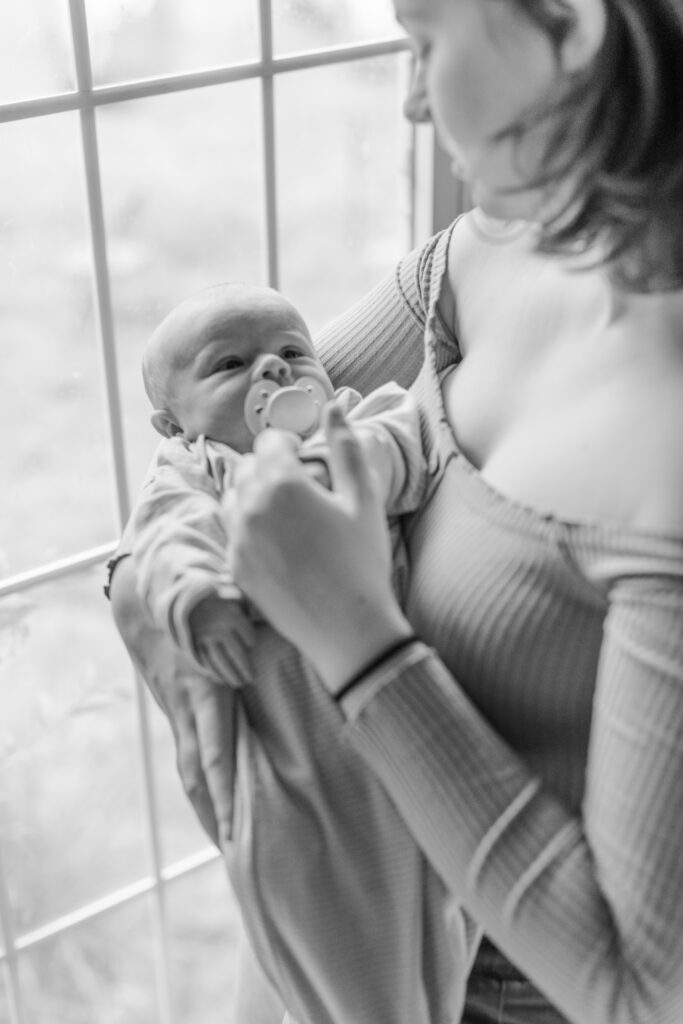 A mother standing in front of a window, gently holding her newborn in her arms, with their hands touching as they look at each other in black and white.