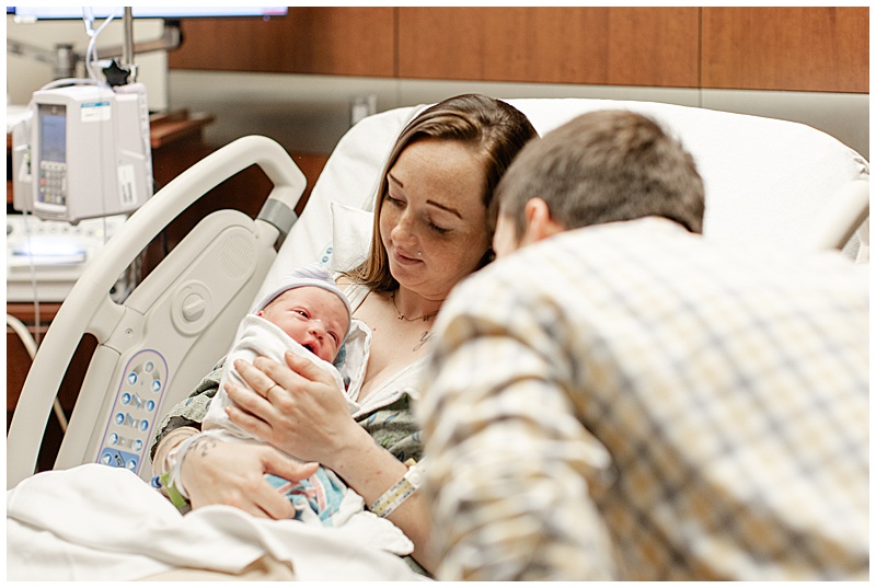 Mother is showing Father, their new newborn who is swaddled in hospital blanket. Newborn's eyes are open looking at father.
