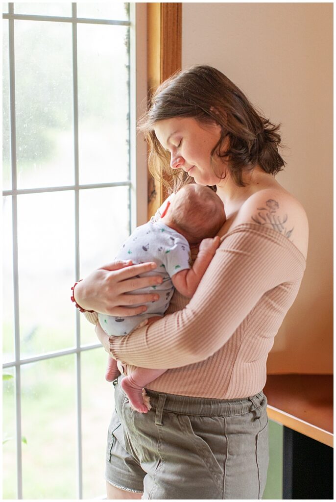 Mother gently holding newborn against chest, standing in front of a large window, looking down at baby