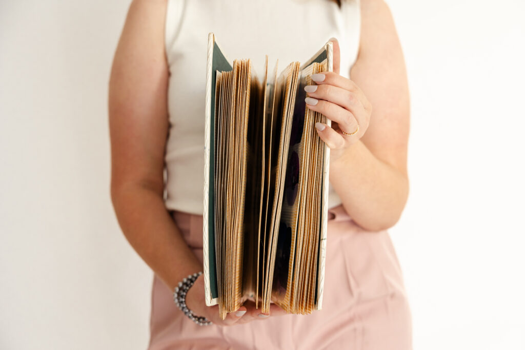 Photographer, Aspin McKenzie Holding a printed photo album against torso, flipping its pages towards the camera.