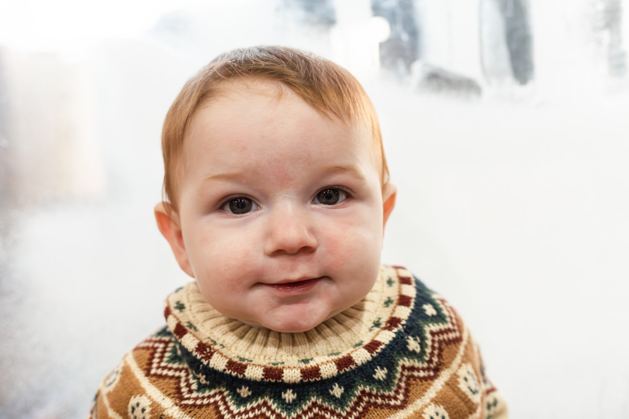 6 month old in a winter sweater smiling at camera in front of frosted window