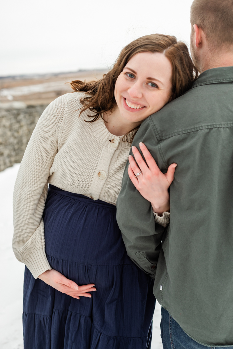 Maternity Photography session, Father has his back to camera while Mother loops his arm and rests her head on his shoulder while she holds baby bump. Wearing blue dress with cream cardigan and green jacket.