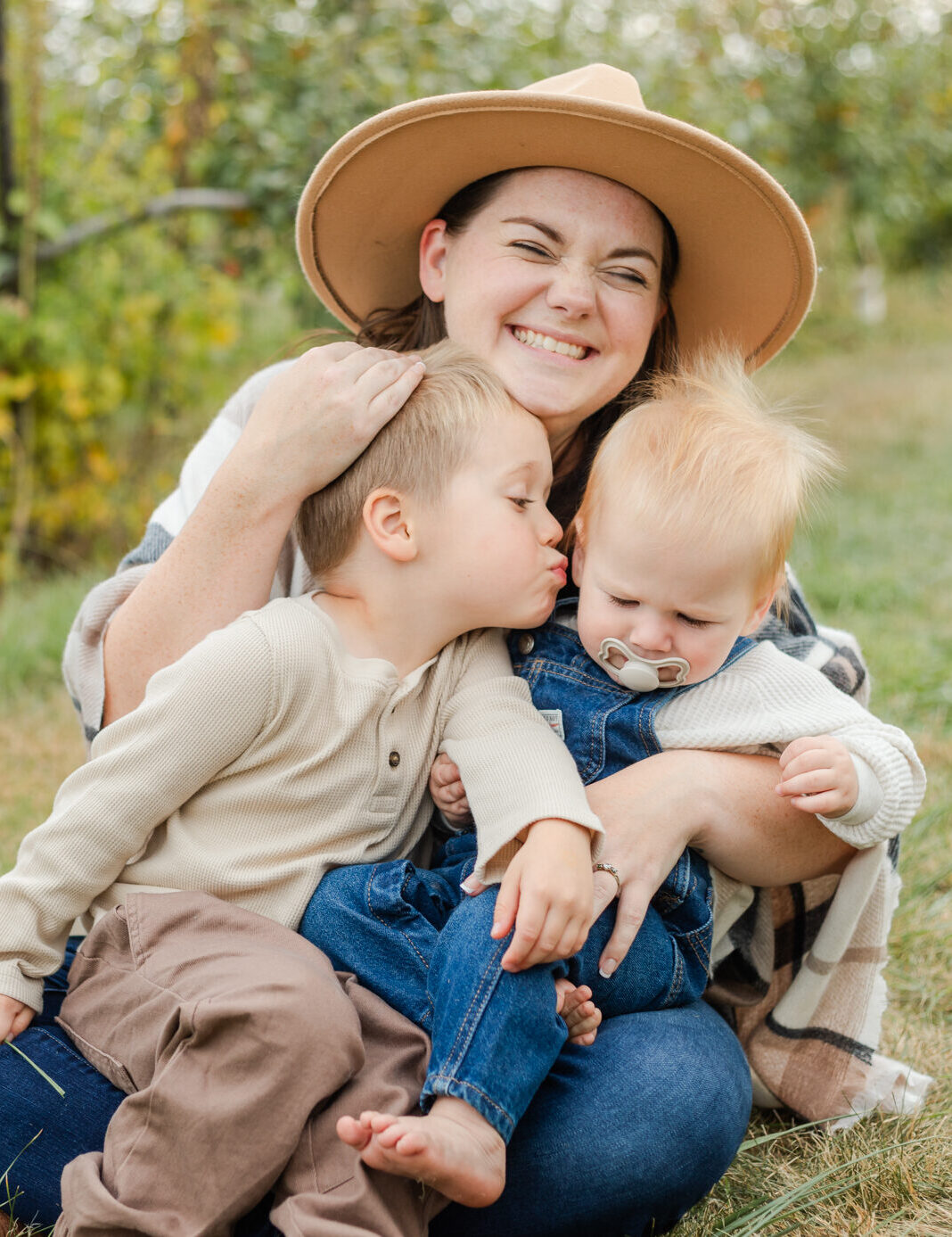 mother holding two boys in her lap smiling, holding the head of the older child while he tries to kiss the infant